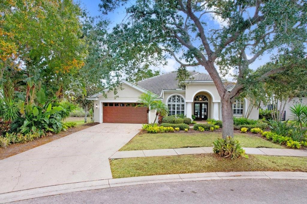 A single-story house with a tan stucco exterior, a brown garage door, and mature trees — 1161 MARINA DRIVE, TARPON SPRINGS, FL