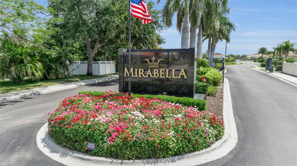 A black monument sign for Mirabella stands behind a circular flower bed with red flowers — 7004 VISTA BELLA DRIVE, BRADENTON, FL
