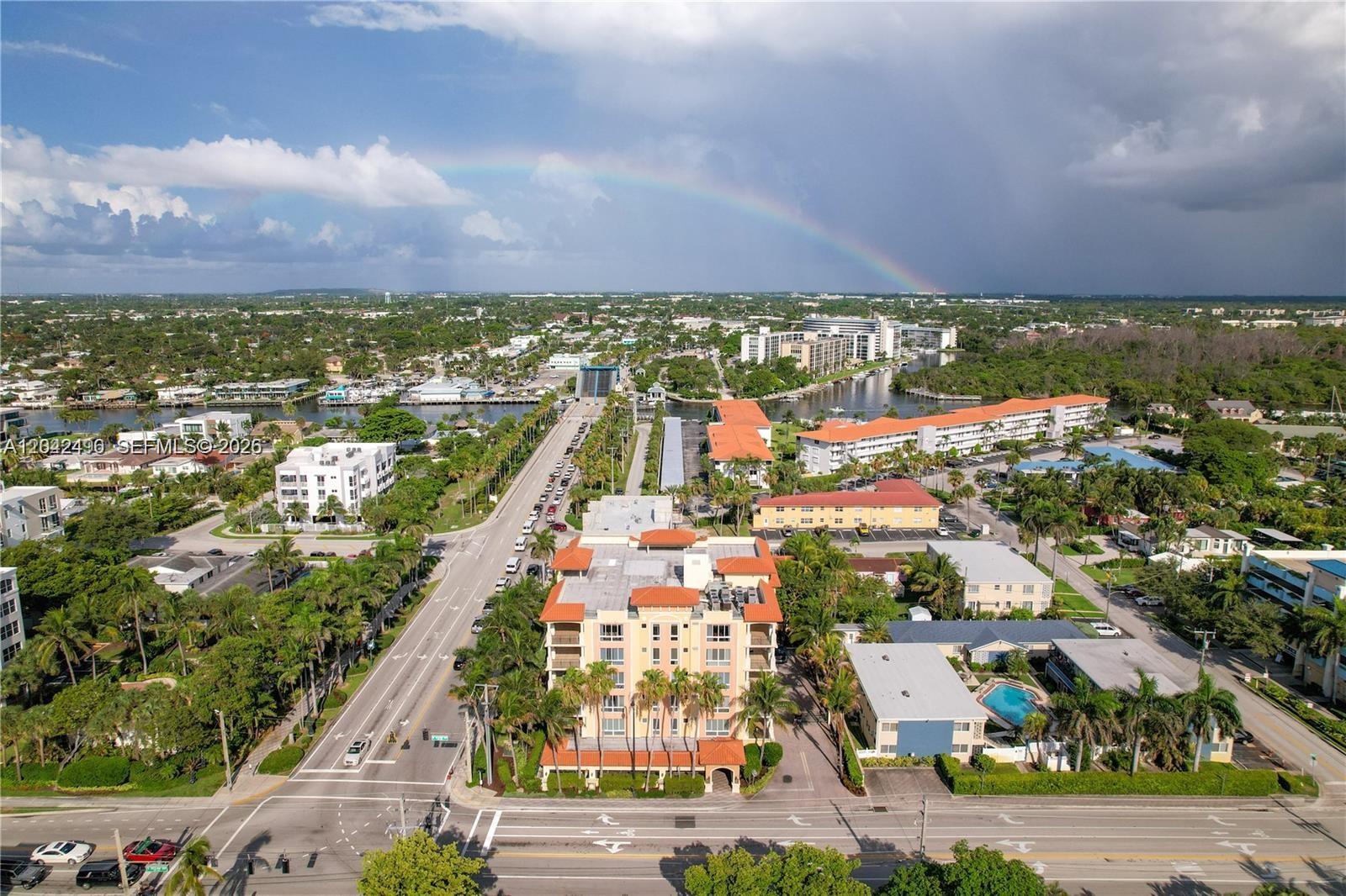 An aerial view shows a multi-story apartment building surrounded by palm trees and a rainbow — 9 NE 20th Ave # 201, Deerfield Beach FL 33441, DEERFIELD BEACH, FL