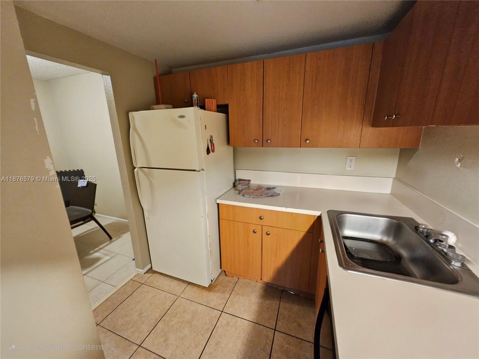 A kitchen features wood-tone cabinets, a white refrigerator, a stainless steel sink, and tiled flooring — 411 Executive Center Dr # 209, West Palm Beach FL 33401, WEST PALM BEACH, FL