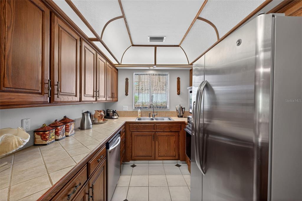 A galley kitchen features wood cabinets, tile countertops, stainless steel appliances, and a vaulted ceiling — 6072 HUNTINGTON WOODS DRIVE UNIT 17, NAPLES, FL