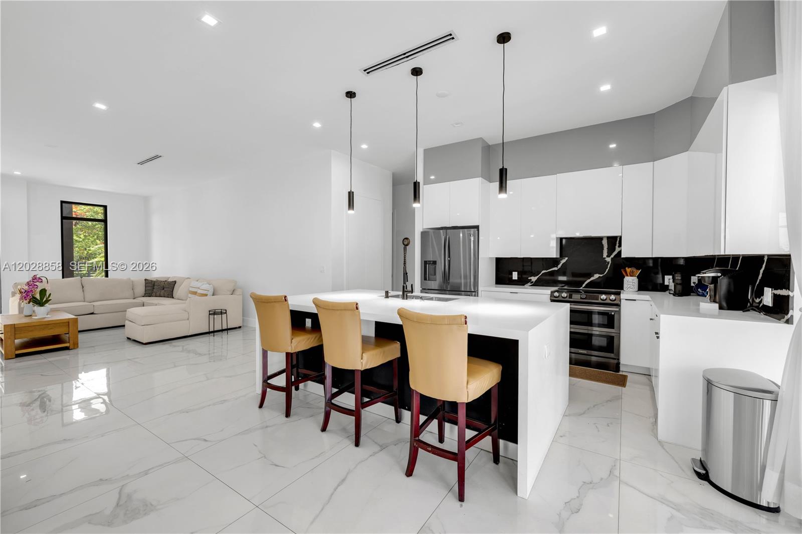 A modern kitchen with white cabinets, a marble island, and bar stools faces a living area — 152 NW 165th St, Miami FL 33169, MIAMI, FL