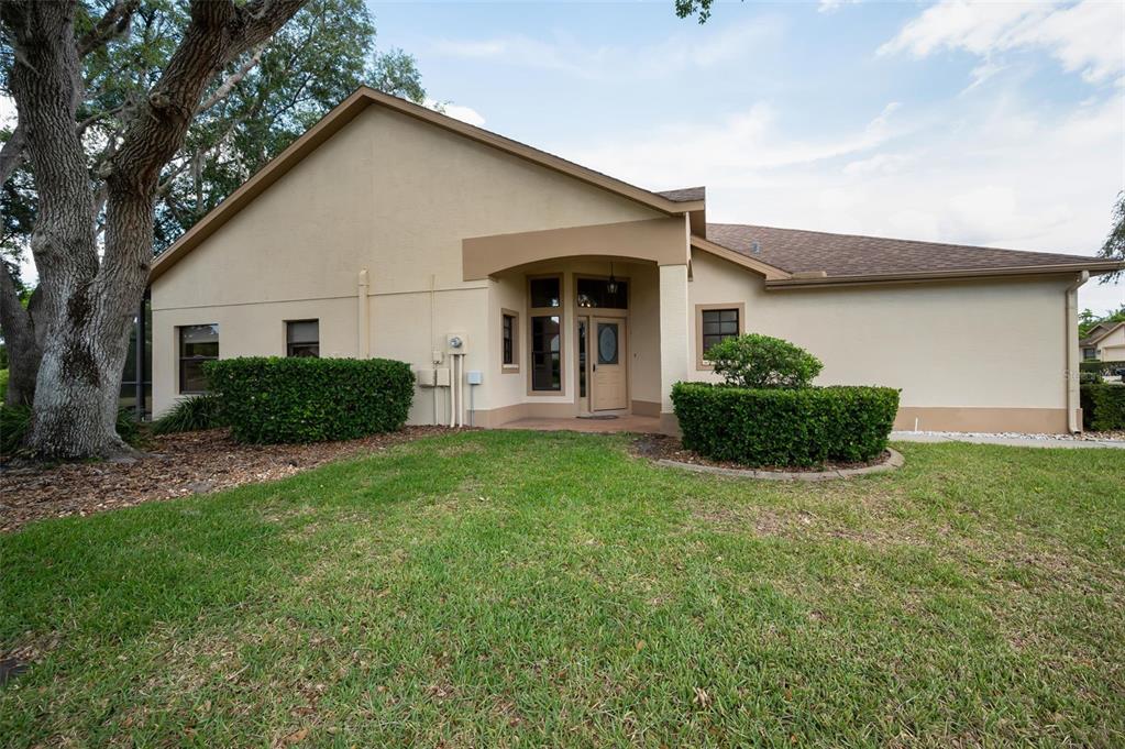 A tan single-story house exterior features a recessed entryway, a large oak tree, and landscaping — 9307 PALM HAVEN COURT, NEW PORT RICHEY, FL