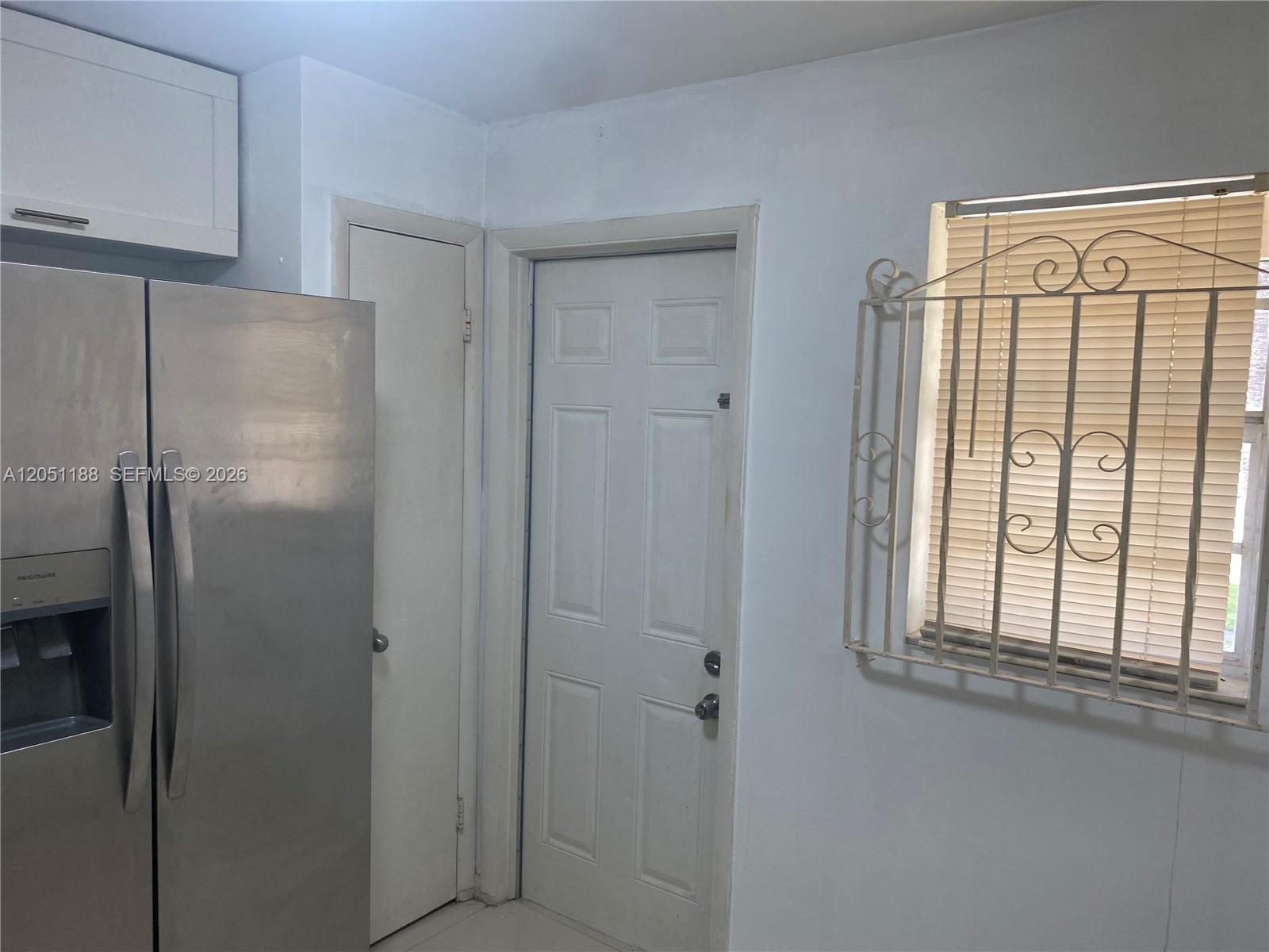 A stainless steel refrigerator stands next to a white door and a window with security bars — 1550 NE 191st St # 309, Miami FL 33179, MIAMI, FL