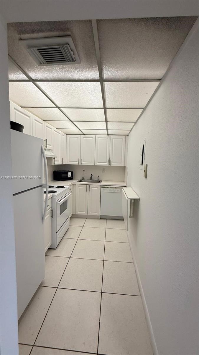 A narrow kitchen features white cabinets, white appliances, tiled floors, and a drop-ceiling grid — 17530 NW 68th Ave # C3010, Hialeah FL 33015, HIALEAH, FL