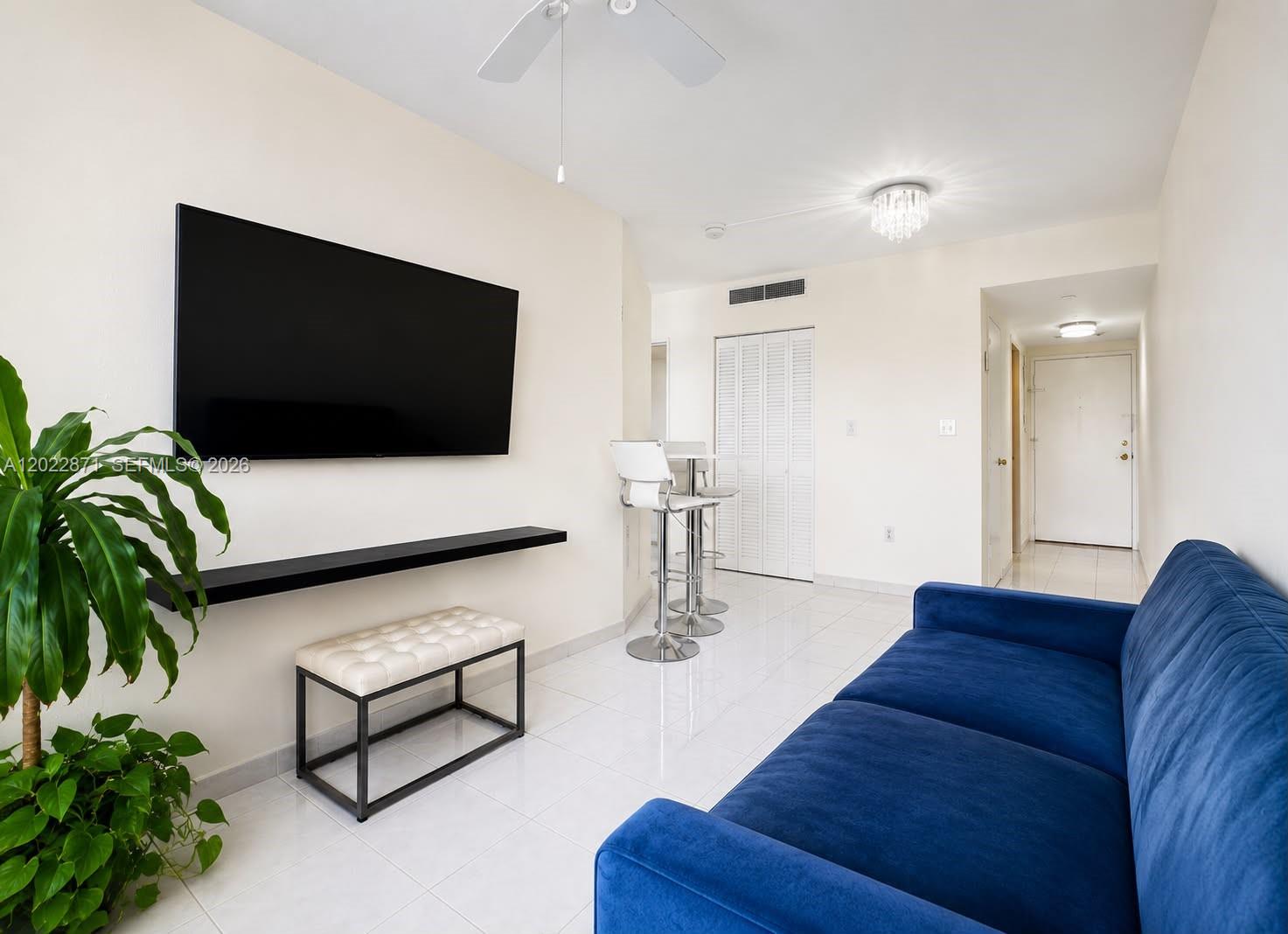 A living room features a blue sofa, wall-mounted television, floating shelf, and white tile floors — 2899 Collins Ave # 1042, Miami Beach FL 33140, MIAMI BEACH, FL