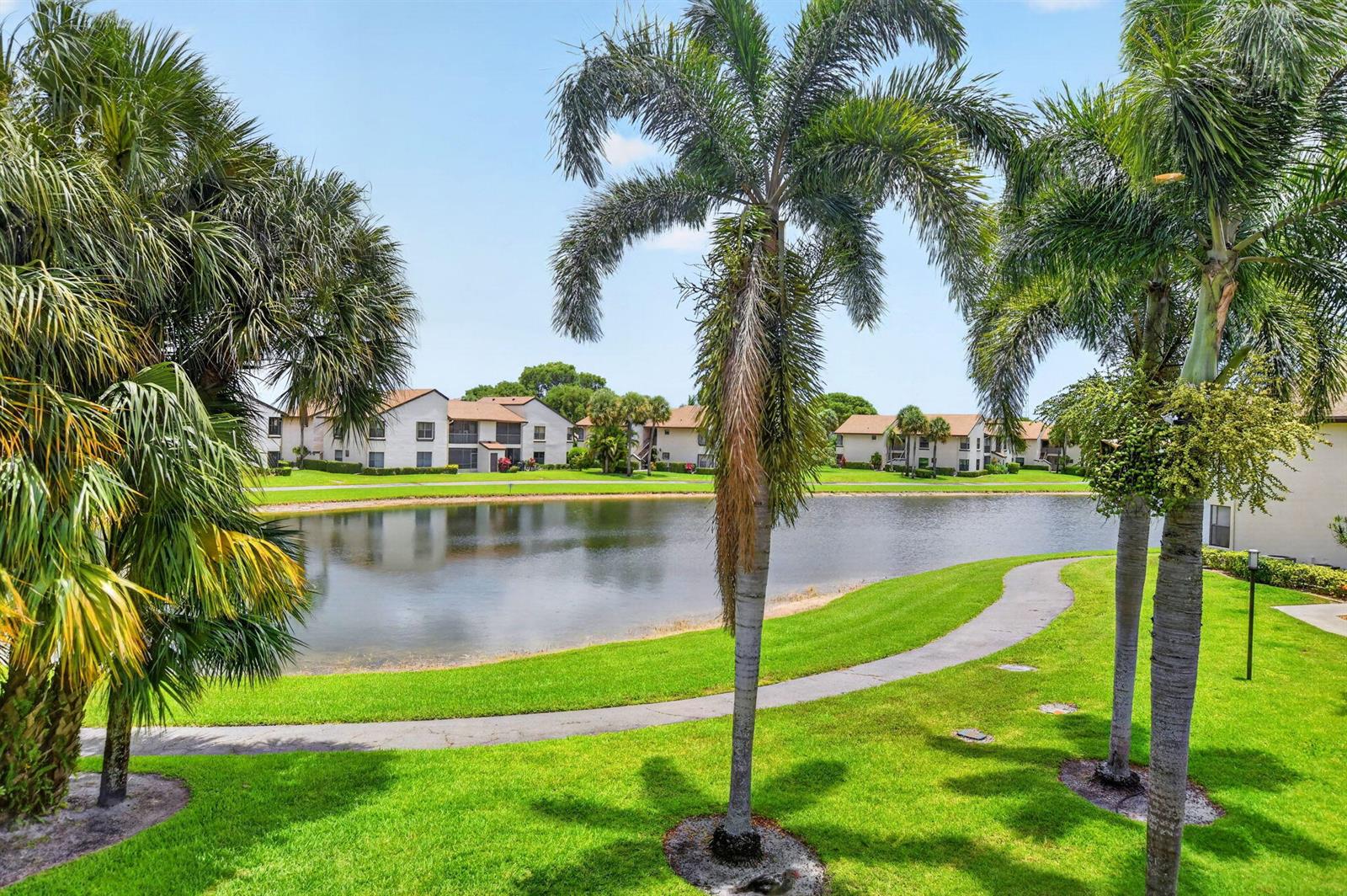 A view of a pond surrounded by green grass, palm trees, and multi-story apartment buildings — 8617 W Boca Glades Boulevard # H, Boca Raton FL 33434, BOCA RATON, FL