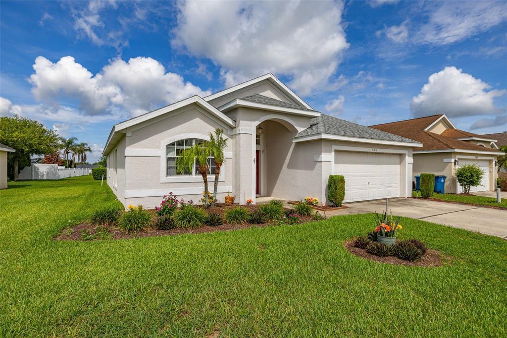A single-story beige house with a two-car garage and a front lawn under a blue sky — 135 JESSAMINE DRIVE, DAVENPORT, FL