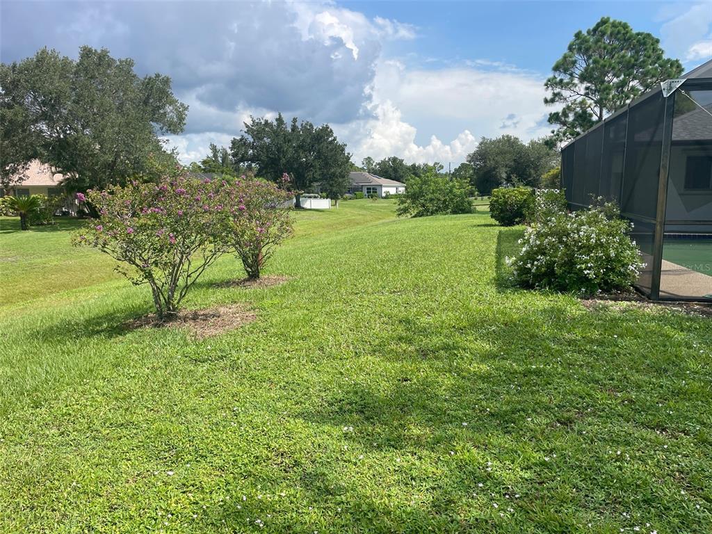 A grassy backyard features two flowering bushes, a pool enclosure, and distant neighboring houses — 1091 VENTURE COURT, PUNTA GORDA, FL