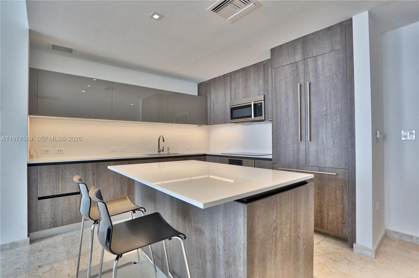 A modern kitchen features wood-grain cabinetry, a white island countertop, and two black chairs — 88 SW 7th St # 3805, Miami FL 33130, MIAMI, FL