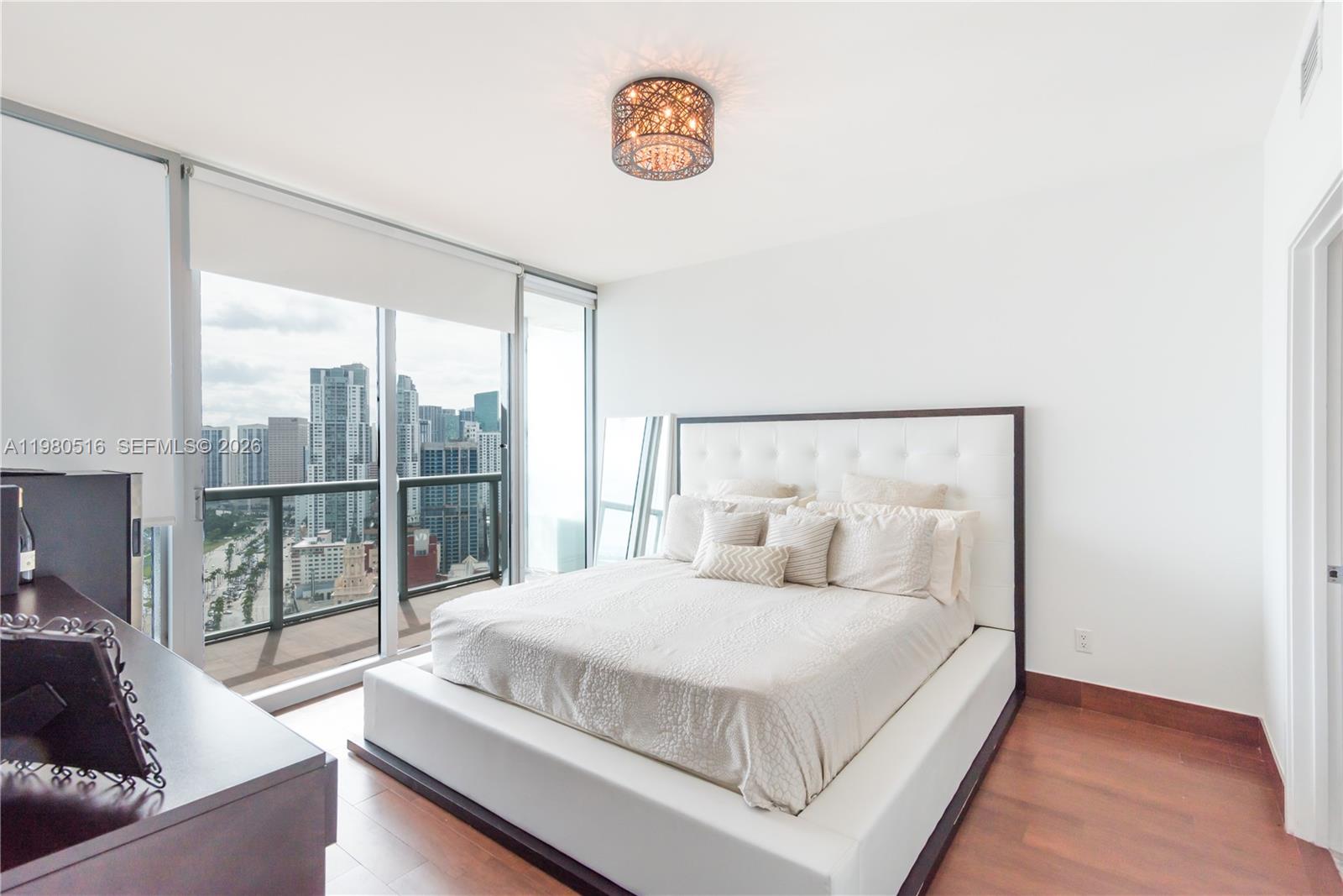 A modern bedroom features a white platform bed, wood flooring, and floor-to-ceiling windows with city views — 888 Biscayne Blvd # 3506, Miami FL 33132, MIAMI, FL