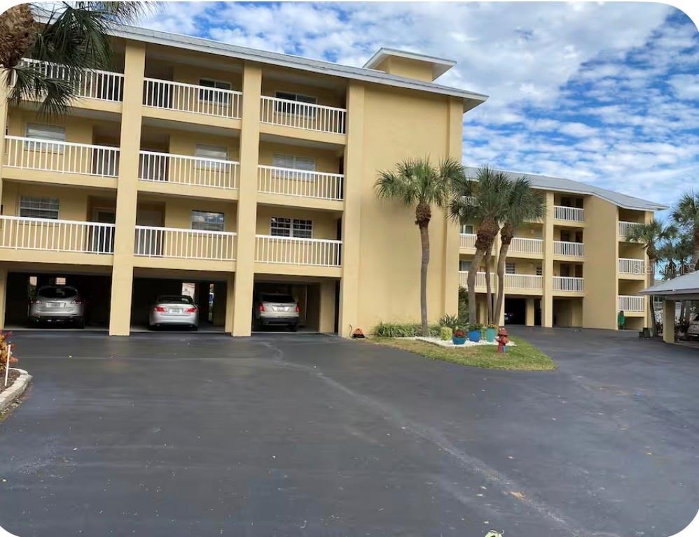 A three-story yellow apartment building with covered ground-level parking and palm trees in front — 1125 PINELLAS BAYWAY S UNIT 201 furnished, ST PETERSBURG, FL