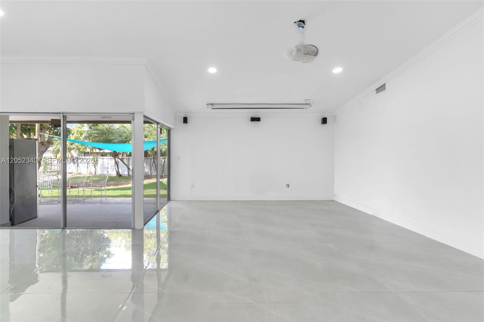 A bright, empty room features large white floor tiles, a projector, and sliding glass doors — 26222 SW 133rd Ct, Homestead FL 33032, HOMESTEAD, FL