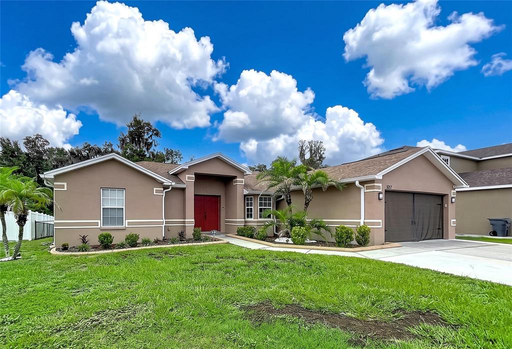 A single-story tan house with a red front door, a garage, and a grassy yard — 3217 ENCLAVE BOULEVARD, MULBERRY, FL