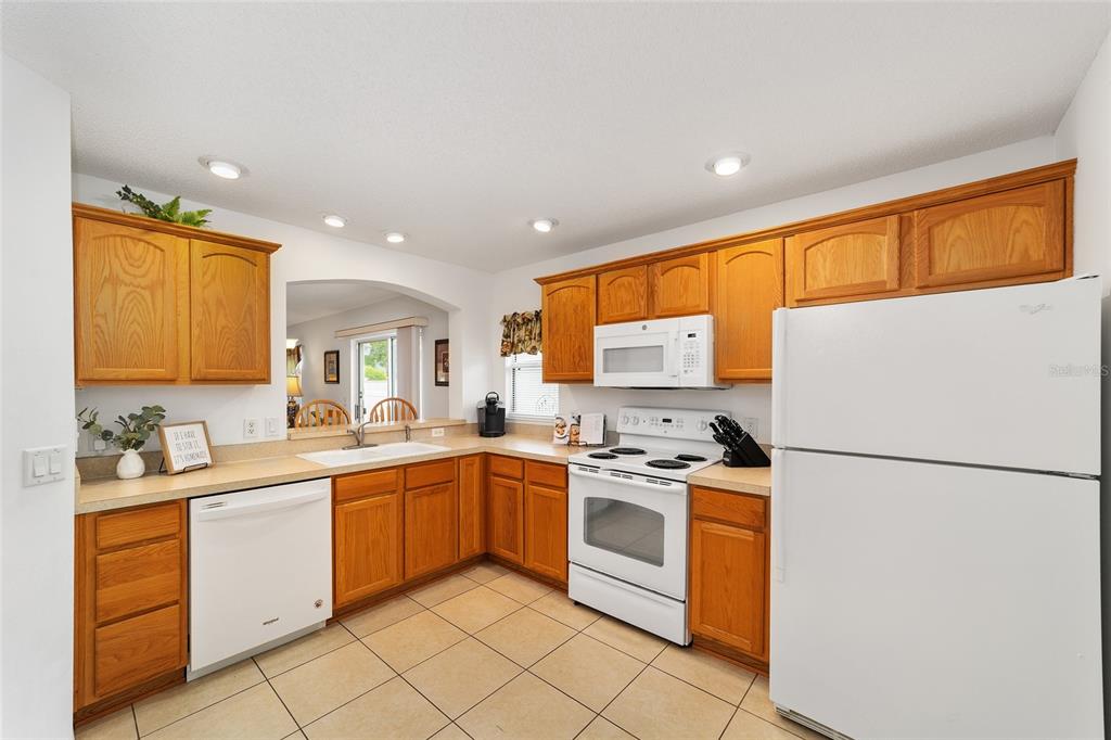 A kitchen with oak cabinets, white appliances, tile flooring, and a pass-through window — 2426 SAFFRON LANE, THE VILLAGES, FL