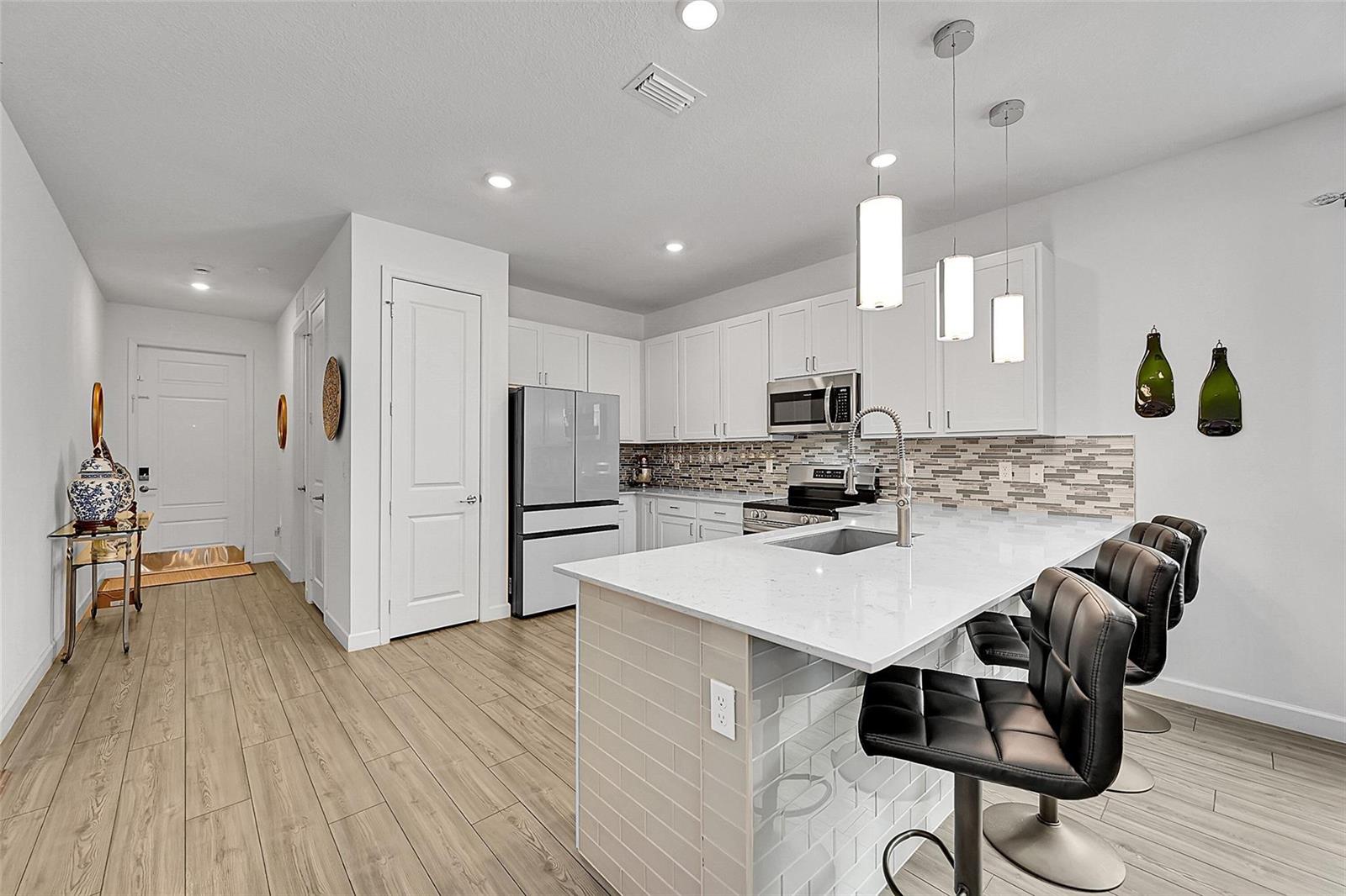 A modern kitchen features white cabinets, quartz countertops, a breakfast bar, and wood-look tile flooring — 13443 Noble Drive, Delray Beach FL 33484, DELRAY BEACH, FL