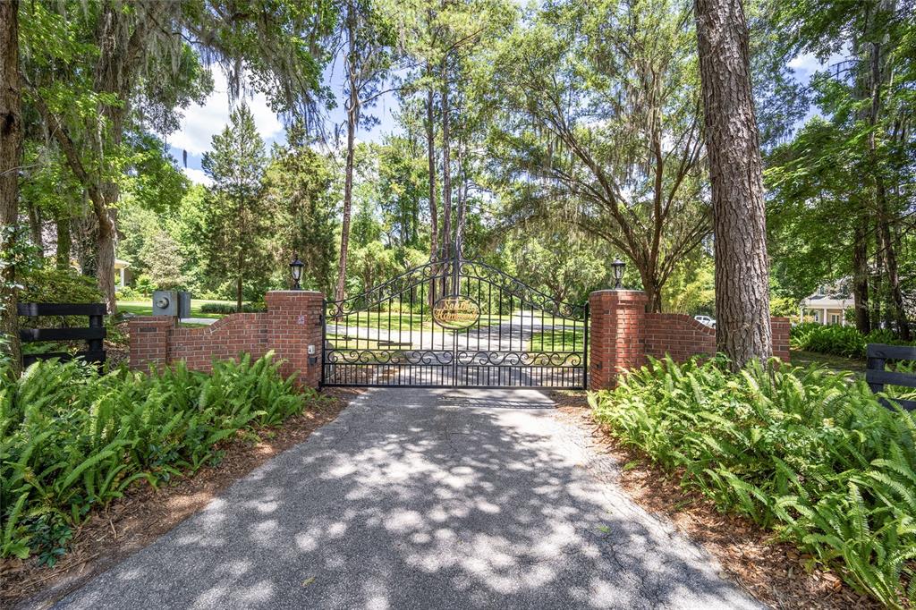 A paved driveway leads to a black wrought iron gate flanked by brick pillars and trees — 4850 SE 11TH AVENUE, OCALA, FL