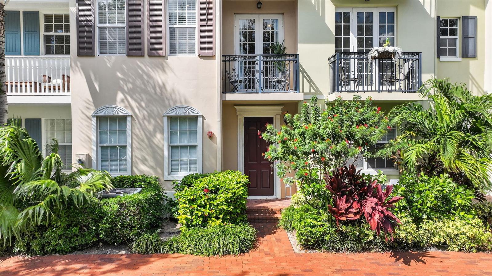 A multi-story townhouse exterior with a brick walkway, lush landscaping, and two upper-level balconies — 1054 E Heritage Club Circle, Delray Beach FL 33483, DELRAY BEACH, FL