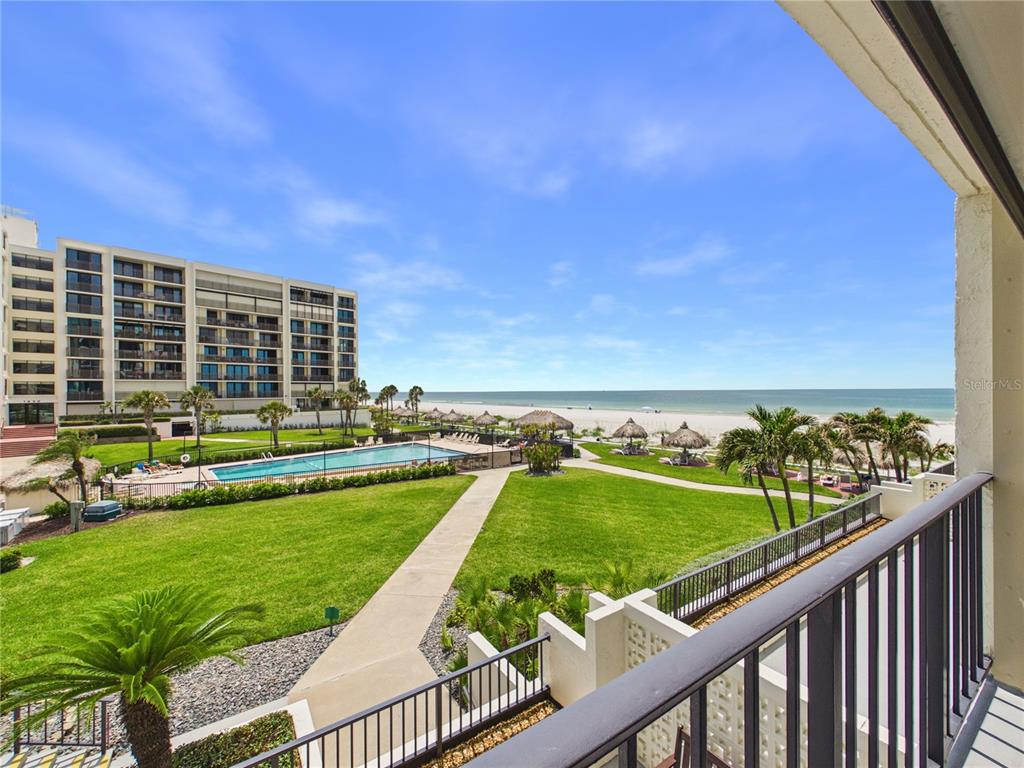 A balcony view overlooks a grassy courtyard, a swimming pool, and the ocean beach — 1400 GULF BOULEVARD UNIT 212, CLEARWATER BEACH, FL