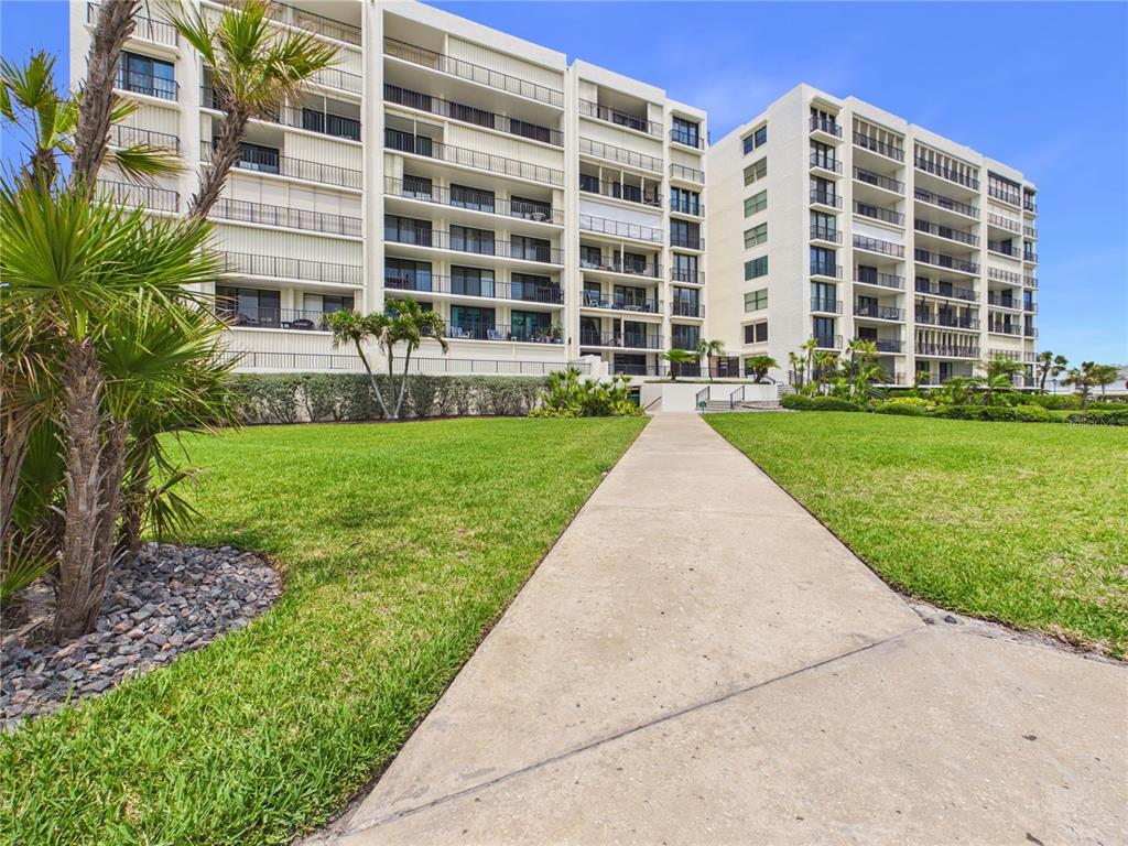 A concrete walkway leads through a grassy lawn toward a multi-story white apartment building — 1400 GULF BOULEVARD UNIT 212, CLEARWATER BEACH, FL