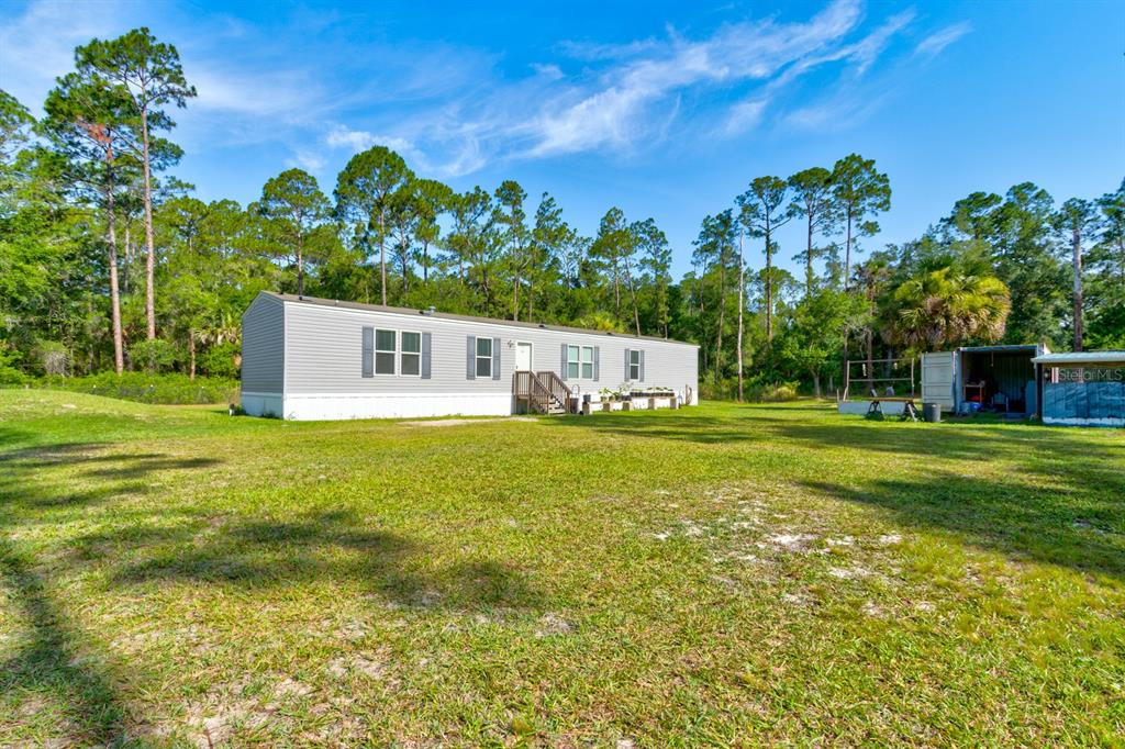 A light-colored manufactured home sits on a large grassy lot surrounded by pine trees — 5074 BUTTERNUT AVENUE, BUNNELL, FL