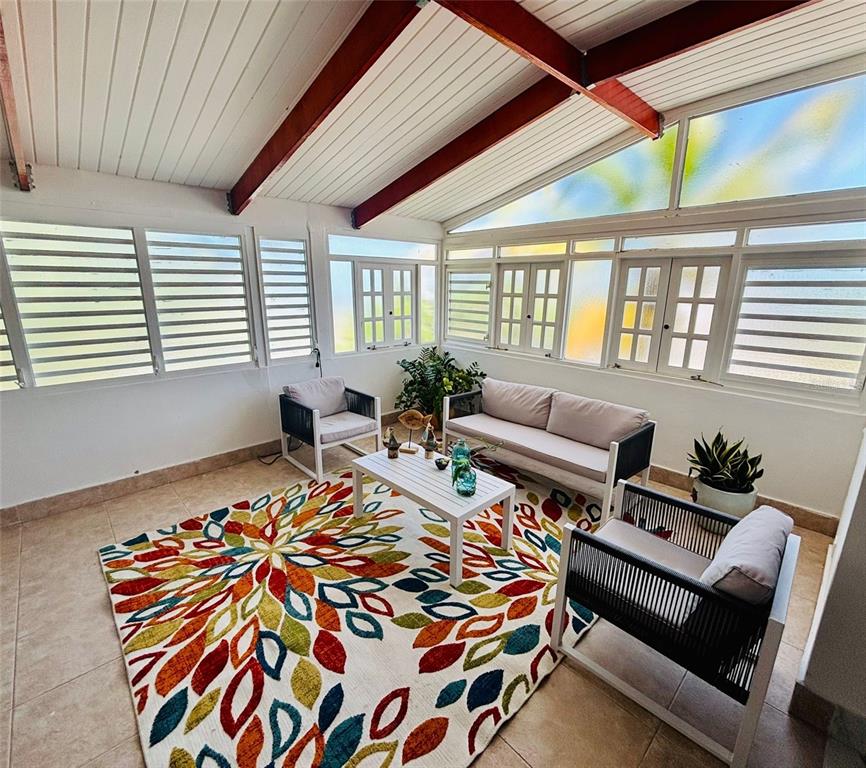 A sunroom features a colorful leaf-patterned rug, white patio furniture, and large windows — Punta Las Marias CAOBA 10 STREET UNIT 3, SAN JUAN, PR