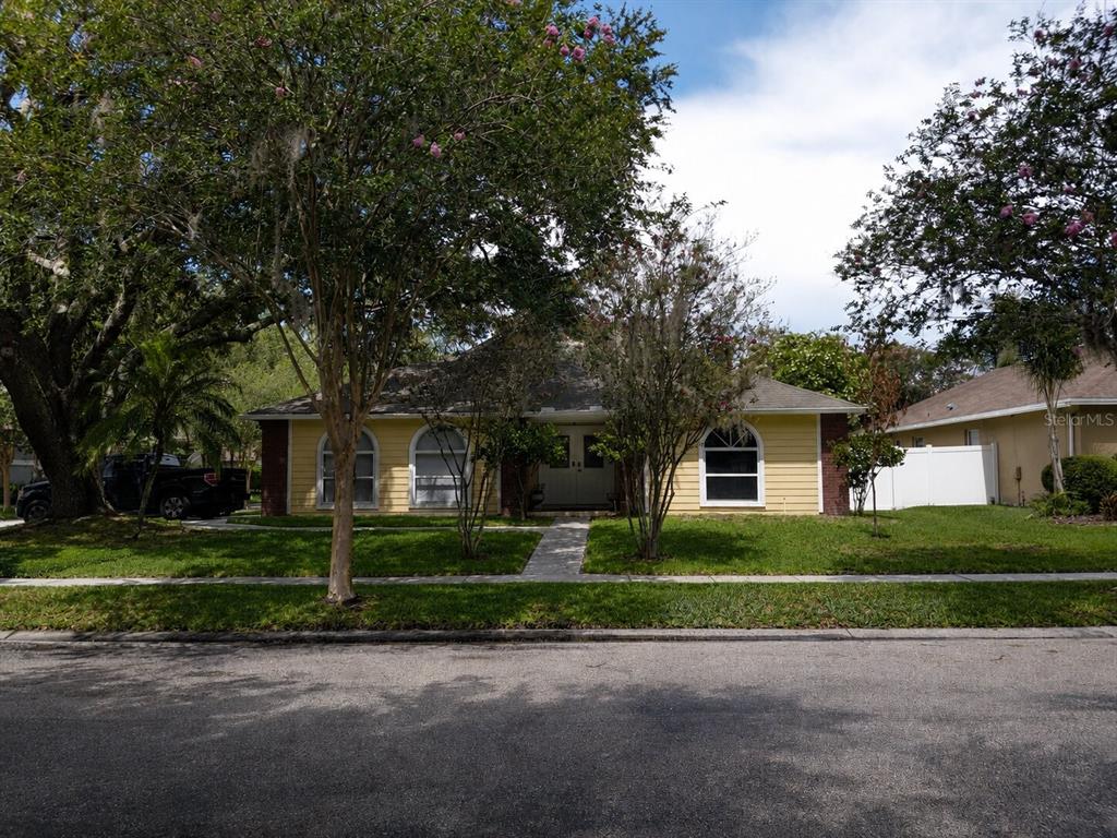 A single-story yellow house with arched windows and a brick-accented entryway sits behind trees — 17502 WOODTHRUSH PLACE, TAMPA, FL