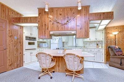 A kitchen features wood-paneled walls, white cabinetry, a central island with two chairs, and carpeting — 35909 BUENO DRIVE, ZEPHYRHILLS, FL