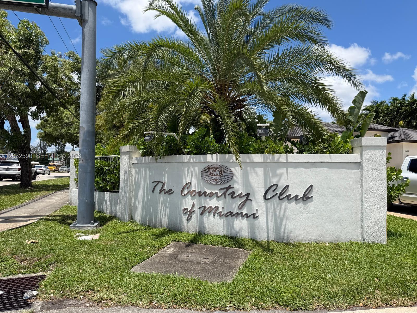 A white stucco entrance sign for The Country Club of Miami sits beneath a palm tree — 18801 Oakland Hills Dr, Hialeah FL 33015, HIALEAH, FL