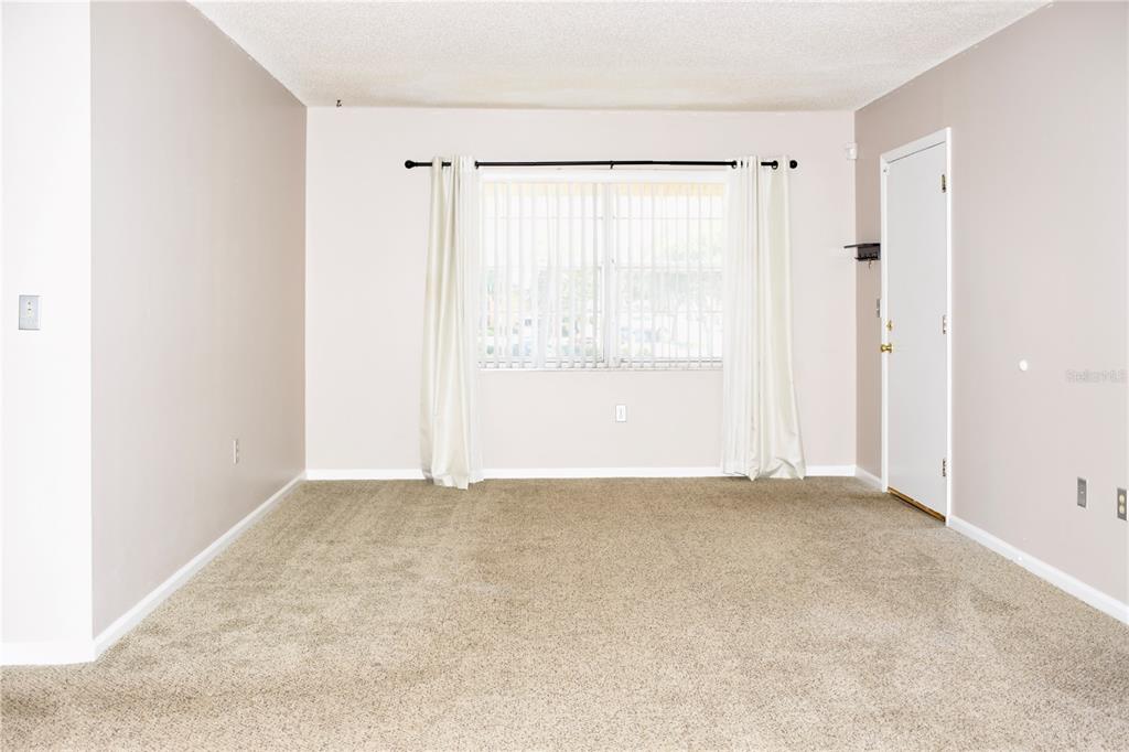 An empty living room features beige carpet, light-colored walls, a window with curtains, and a door — 1920 E EDGEWOOD DRIVE UNIT A8, LAKELAND, FL
