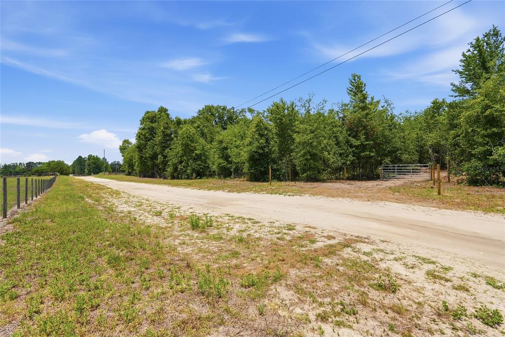 A dirt road runs alongside a fenced, wooded lot with a metal gate under blue skies — SW 188TH ST, ARCHER, FL