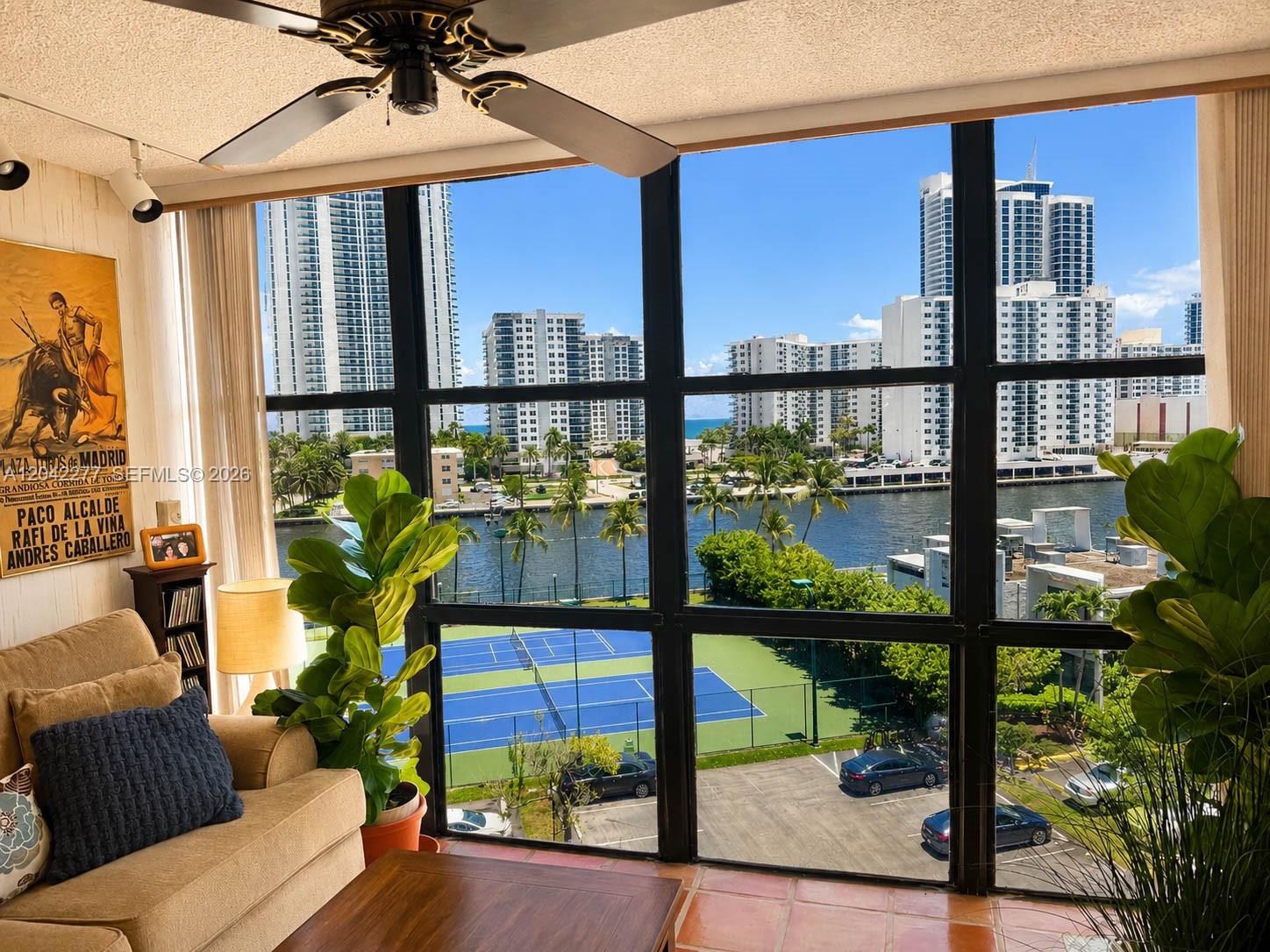 A living room with a large window overlooking a tennis court, canal, and city skyline — 1000 Parkview Dr # 726, Hallandale Beach FL 33009, HALLANDALE BEACH, FL