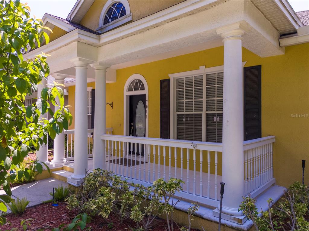 A yellow house exterior features a covered front porch with white columns and railings — 7011 NOBLETON DRIVE, WINDERMERE, FL