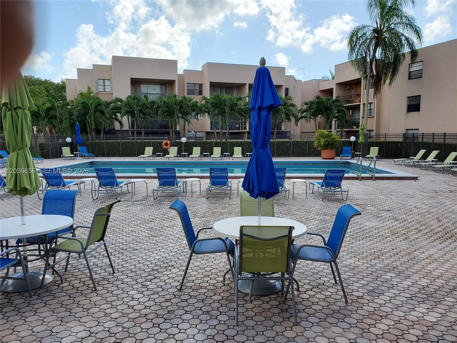 A swimming pool area features lounge chairs, patio tables with umbrellas, and multi-story apartment buildings — 10425 SW 112th Ave # 207, Miami FL 33176, MIAMI, FL