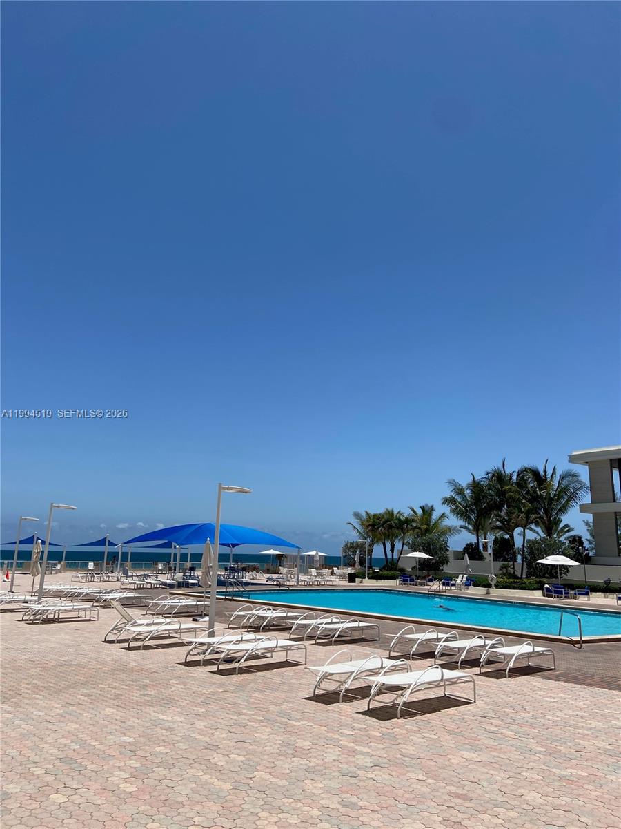 A swimming pool with lounge chairs and blue shade structures sits by the ocean — 3180 S Ocean Dr # 718, Hallandale Beach FL 33009, HALLANDALE BEACH, FL