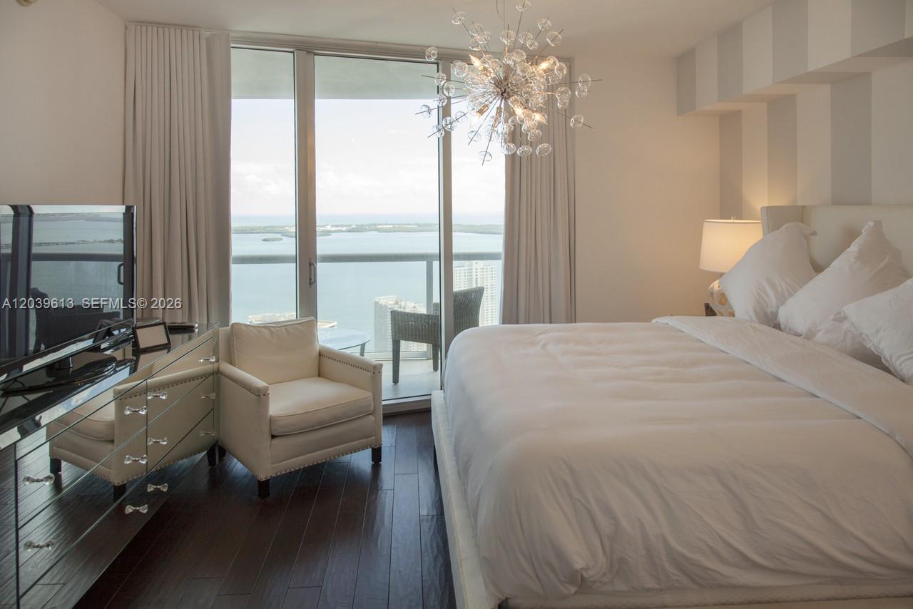 A bedroom features a bed, armchair, mirrored dresser, and floor-to-ceiling windows overlooking the ocean — 465 Brickell Ave # 4703, Miami FL 33131, MIAMI, FL