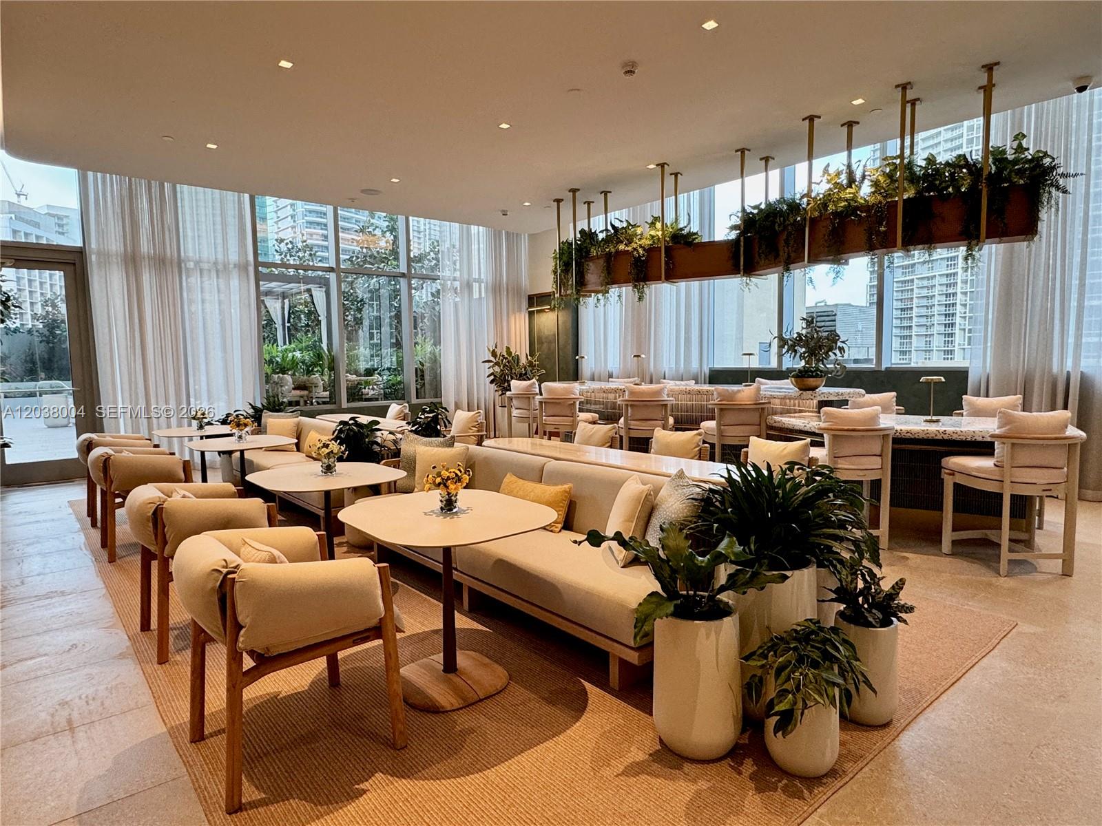 A bright lounge area features modern furniture, potted plants, and large windows with sheer curtains — 77 SE 5th St # 4002, Miami FL 33131, MIAMI, FL