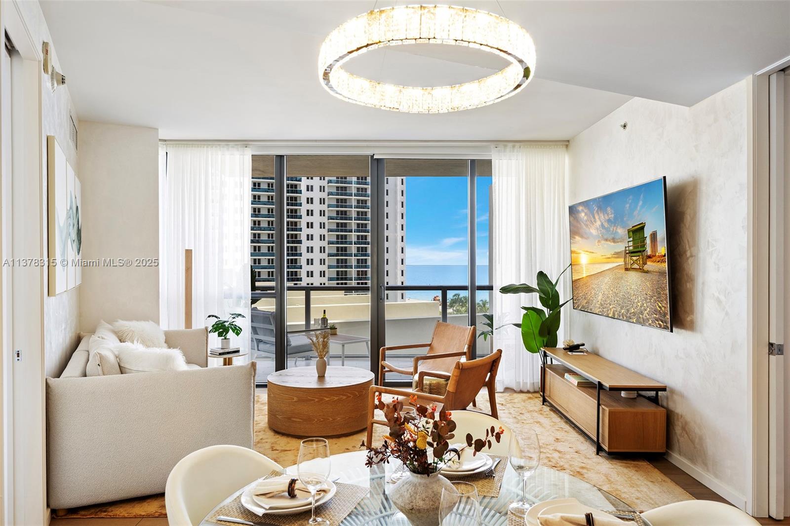 A living room features a circular chandelier, a beige sofa, wooden chairs, and a balcony view — 2201 Collins Ave # 811, Miami Beach FL 33139, MIAMI BEACH, FL
