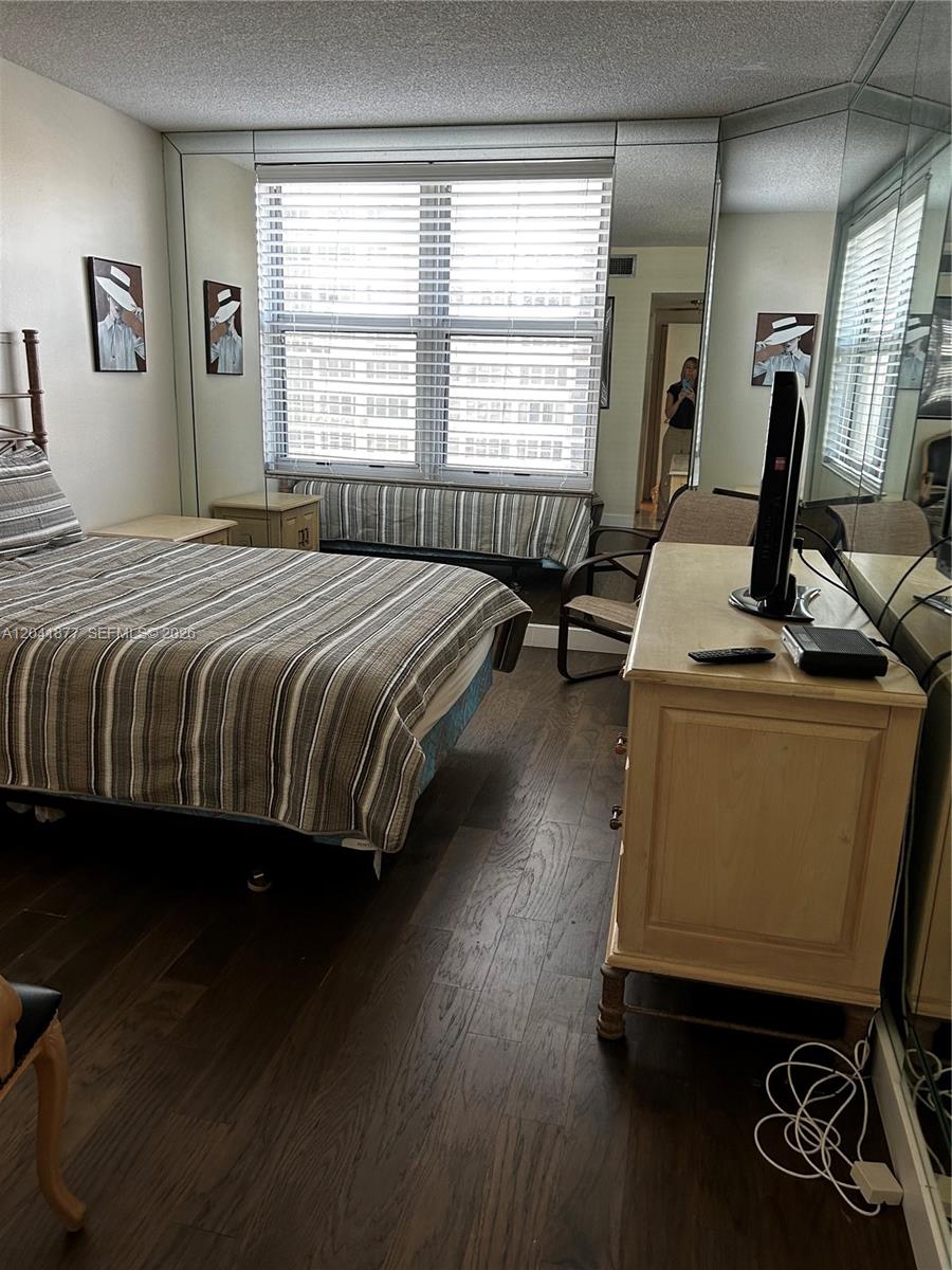 A bedroom features a striped bedspread, wood flooring, mirrored walls, and a window with blinds — 1980 S Ocean Dr # 8A, Hallandale Beach FL 33009, HALLANDALE BEACH, FL