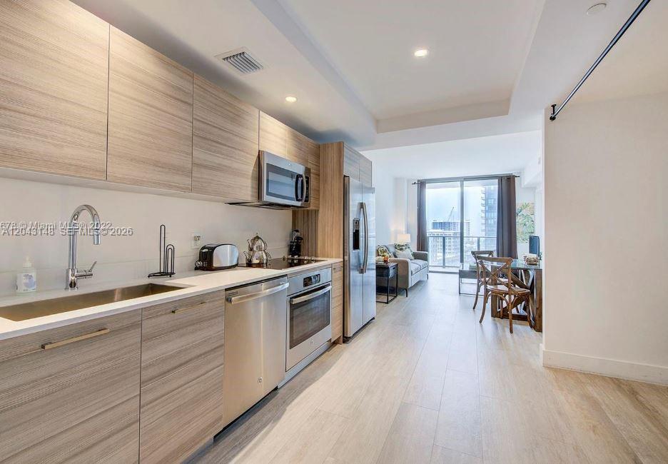 A modern kitchen with light wood cabinets and stainless steel appliances opens into a living area — 121 NE 34th St # 1603, Miami FL 33137, MIAMI, FL