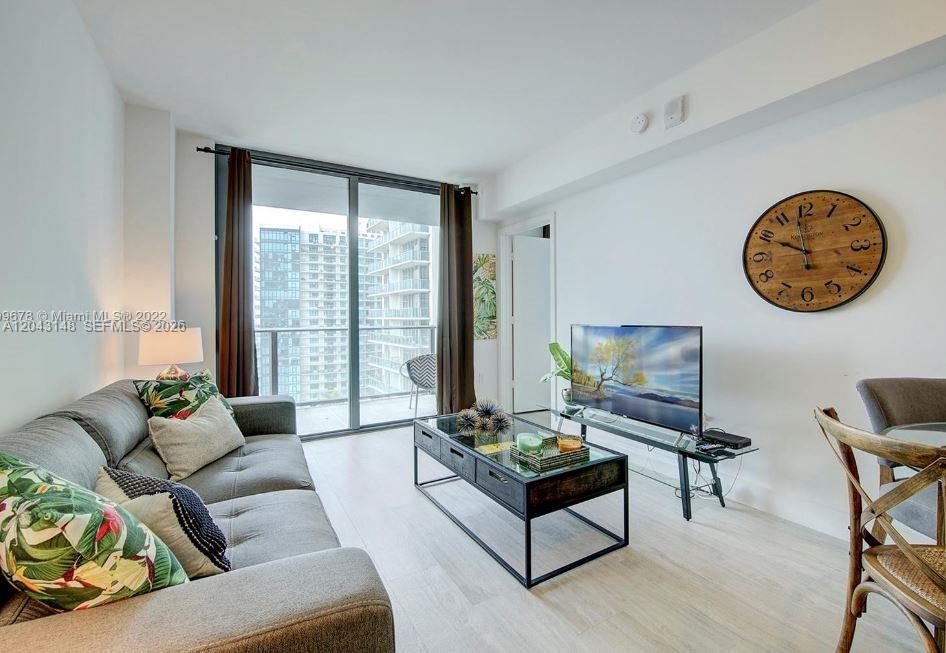 A living room features a gray sofa, glass coffee table, television, and a large wall clock — 121 NE 34th St # 1603, Miami FL 33137, MIAMI, FL