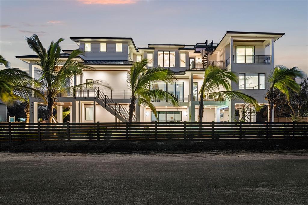A modern three-story house with palm trees, a wooden fence, and an exterior staircase at dusk — 7007 POINT OF ROCKS ROAD, SARASOTA, FL
