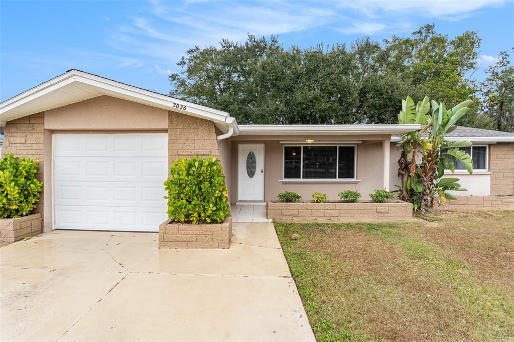A single-story house with a white garage door, tan masonry, and a front garden bed — 3036 BLUE BIRD DRIVE, HOLIDAY, FL