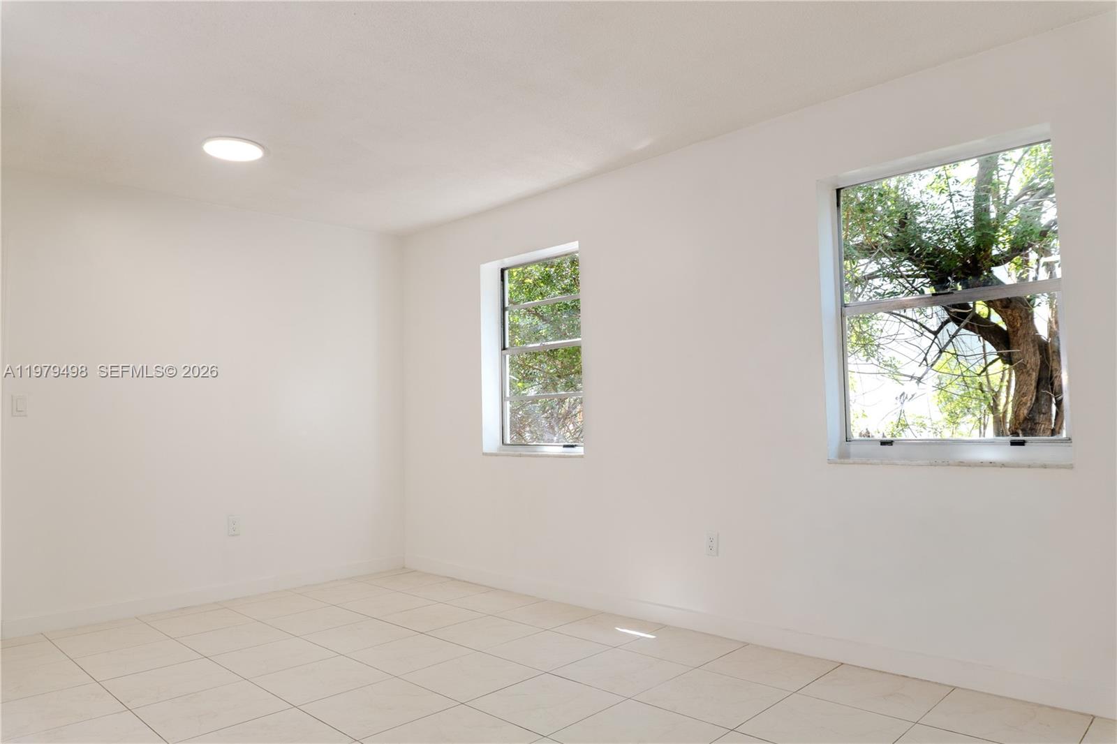 An empty room features white walls, light-colored tile flooring, and two windows overlooking trees — 1525 NW 19th Ter # 15, Miami FL 33125, MIAMI, FL