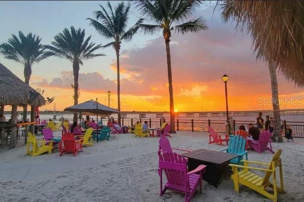 A sandy outdoor patio features colorful Adirondack chairs, palm trees, and a sunset over water — 21471 QUESADA AVENUE, PORT CHARLOTTE, FL