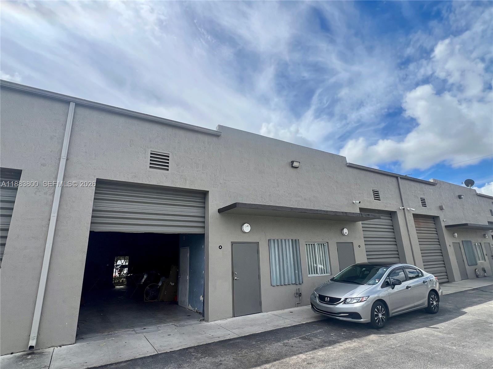 An exterior view of a commercial warehouse building with roll-up doors and a parked car — 16215 SW 117th Ave # 4, Miami FL 33177, MIAMI, FL