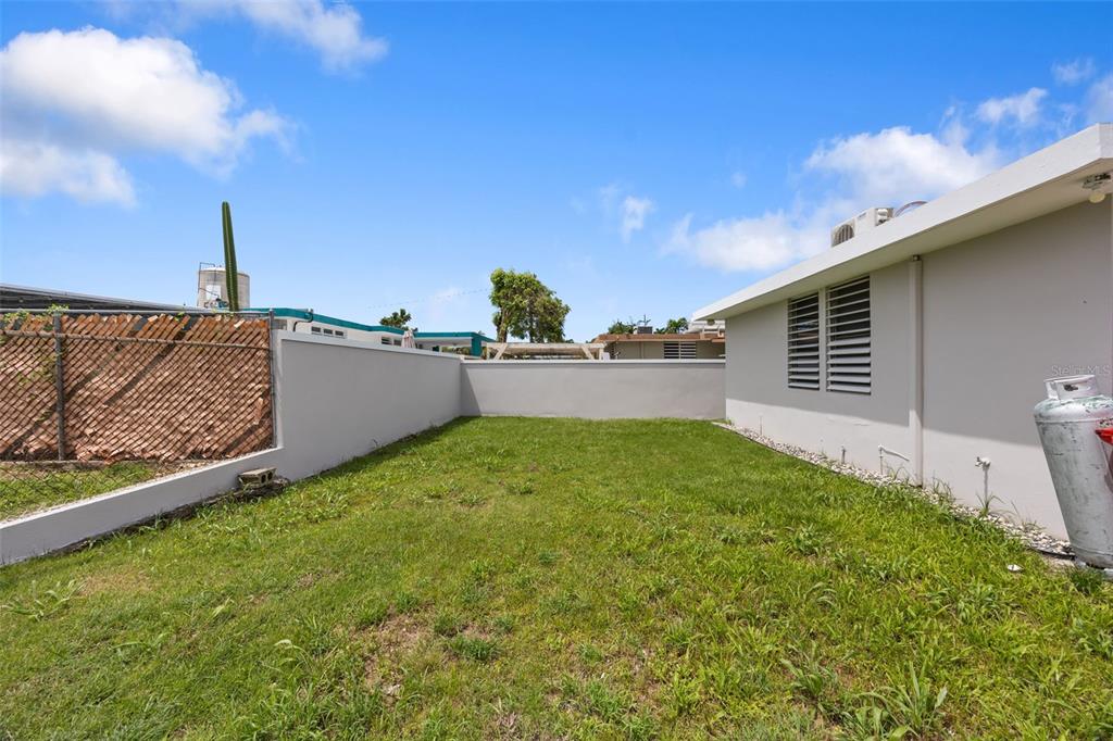A grassy backyard area features a concrete wall, a chain-link fence, and a house exterior — CASA SUAVE CALLE 4 UNIT B-26, AGUADA, PR