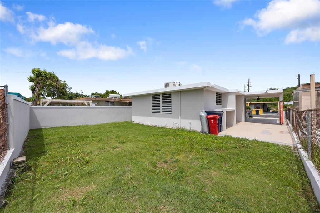 A grassy backyard features a light-colored house with a covered patio and a perimeter fence — CASA SUAVE CALLE 4 UNIT B-26, AGUADA, PR