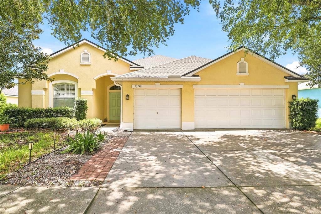 A yellow single-story house features a three-car garage, a brick walkway, and mature trees — 16748 MIDSUMMER LANE, SPRING HILL, FL