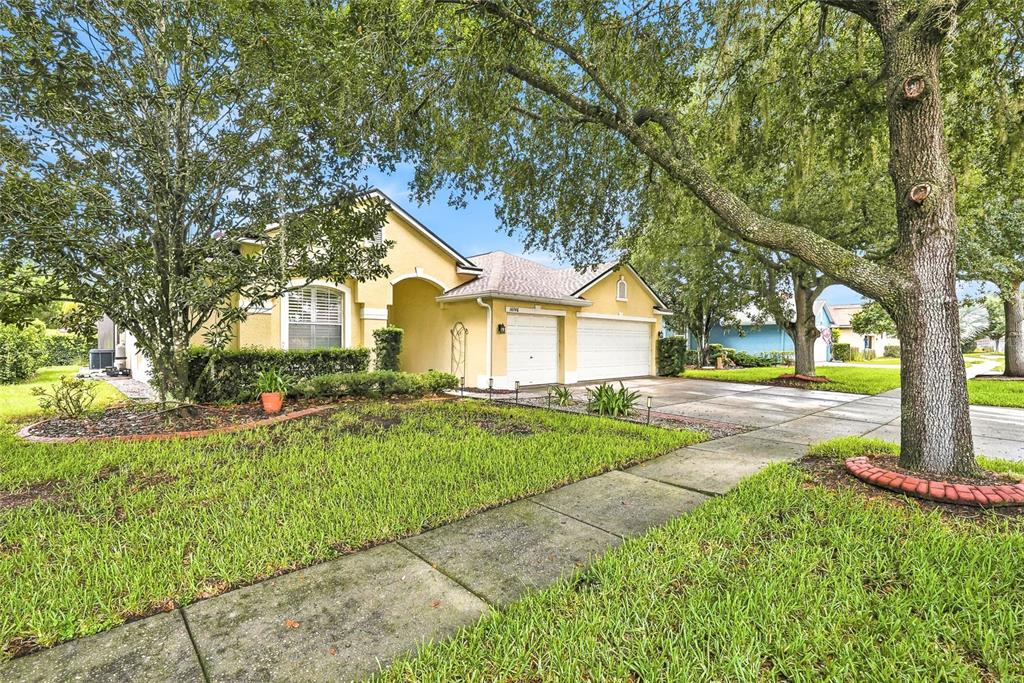 A yellow single-story house with a two-car garage and mature trees in the front yard — 16748 MIDSUMMER LANE, SPRING HILL, FL